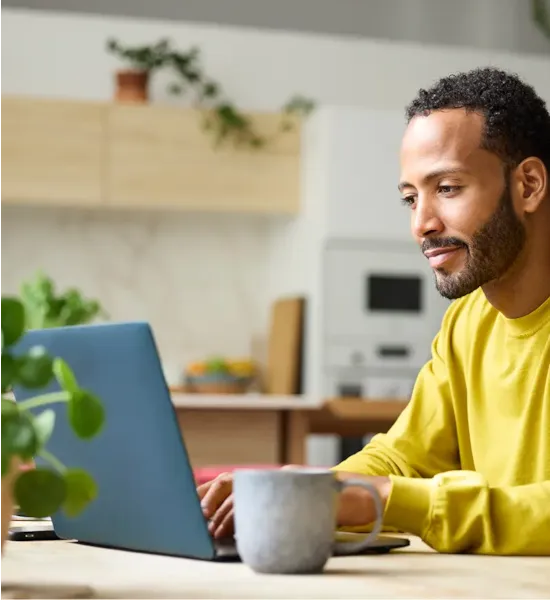 Man in a yellow sweater working on a laptop at a kitchen table with a mug and plants nearby.
